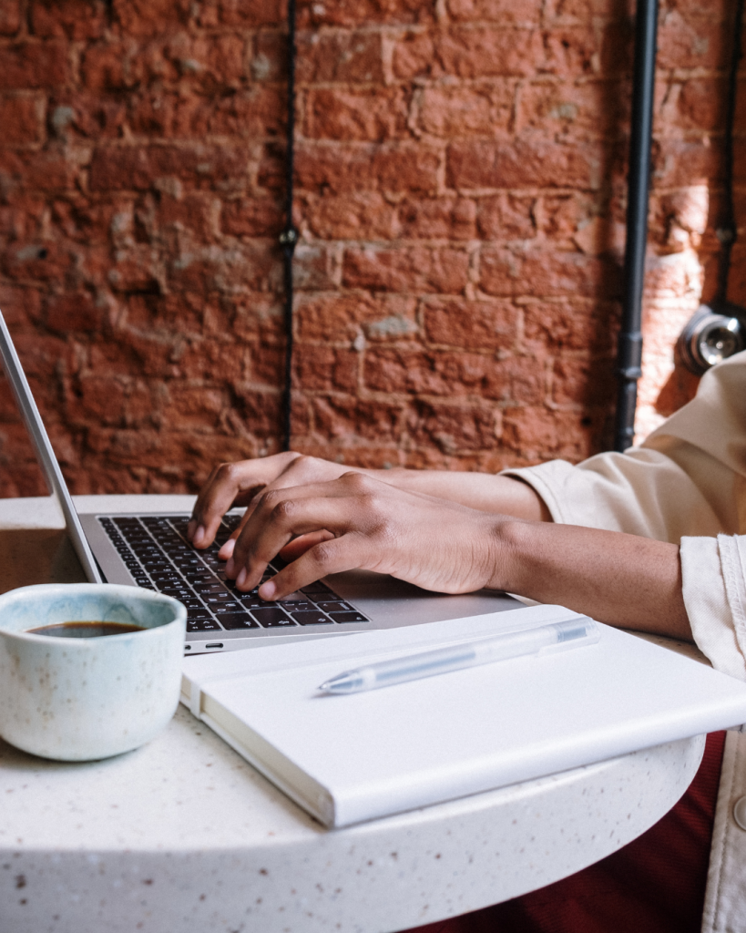 Person typing on a laptop while creating a mailing list, with a notebook, pen, and coffee cup on a round table in front of an exposed brick wall.